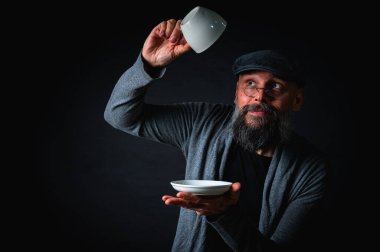 Boş bir fincan ve fincan tabağı tutan düz şapkalı sakallı adam şaşırdı, kaşlarını kaldırarak kontrol ediyor. Komik ifade, karanlık arka planlı stüdyo çekimi.