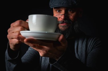 Boş bir fincan ve fincan tabağı tutan düz şapkalı sakallı adam şaşırdı, kaşlarını kaldırarak kontrol ediyor. Komik ifade, karanlık arka planlı stüdyo çekimi.