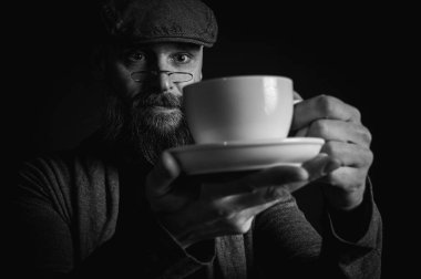 Siyah ve beyaz portre, şık şapkalı ve gözlüklü olgun sakallı bir adam gizemli bir gülümsemeyle bir fincan kahve sunuyor. Olağanüstü aydınlatma ve karanlık arka plan eski moda bir hava yaratıyor..