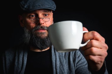 Şık şapkalı ve gözlüklü olgun sakallı bir adam gizemli bir gülümsemeyle bir fincan kahve sunuyor. Dramatik ışıklandırma ve karanlık arkaplan klasik mod yaratır