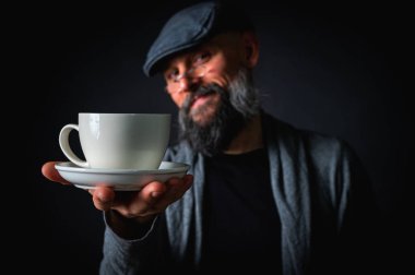 Şık şapkalı ve gözlüklü olgun sakallı bir adam gizemli bir gülümsemeyle bir fincan kahve sunuyor. Dramatik ışıklandırma ve karanlık arkaplan klasik mod yaratır