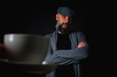 Şık şapkalı ve gözlüklü olgun sakallı bir adam gizemli bir gülümsemeyle bir fincan kahve sunuyor. Dramatik ışıklandırma ve karanlık arkaplan klasik mod yaratır