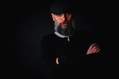 Gözlüklü, düz şapkalı, gölgede oturan düşünceli sakallı adam. Kollarını arkada kavuşturdu. Derin kontrastlı ve karamsar ışıklandırmalı stüdyo portresi