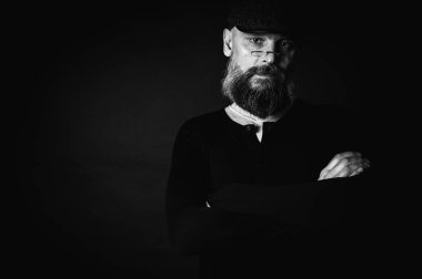 Klasik siyah beyaz fotoğraf, düz şapkalı, siyah arka planda gözlüklü, ciddi sakallı bir adamın yakın çekimi. Şık, zengin görünüşlü, kendine güvenen ve gizemli bir ifade..