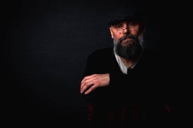 Gözlüklü, düz şapkalı, gölgede oturan düşünceli sakallı adam. Kollarını arkada kavuşturdu. Derin kontrastlı ve karamsar ışıklandırmalı stüdyo portresi