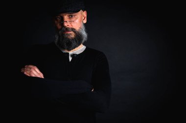 Gözlüklü, düz şapkalı, gölgede oturan düşünceli sakallı adam. Kollarını arkada kavuşturdu. Derin kontrastlı ve karamsar ışıklandırmalı stüdyo portresi