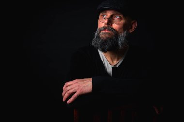 Gözlüklü, düz şapkalı, gölgede oturan düşünceli sakallı adam. Kollarını arkada kavuşturdu. Derin kontrastlı ve karamsar ışıklandırmalı stüdyo portresi