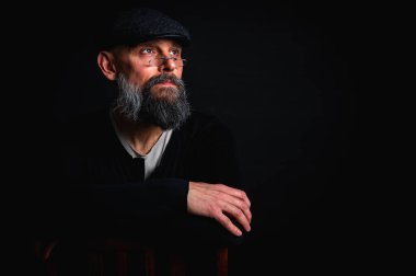 Gözlüklü, düz şapkalı, gölgede oturan düşünceli sakallı adam. Kollarını arkada kavuşturdu. Derin kontrastlı ve karamsar ışıklandırmalı stüdyo portresi