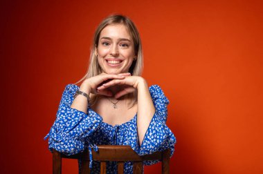 Şaşırmış, duygusal, mavi çiçekli elbiseli, yüzünün yanında ellerini kaldırmış, elleri turuncu arka planda, yana bakan genç bir kadın. Parlak yüz ifadesi, ve neşeli enerji, duygular.