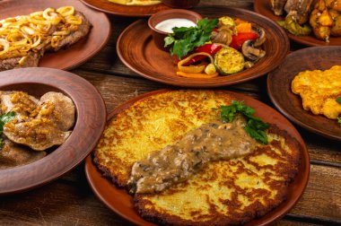 Geleneksel Doğu Avrupa mutfağı kırsal kil tabaklarda servis edilir. Ahşap bir arka planda et, patates, krep, hamur tatlısı ve ızgara sebze gibi ev yapımı yemekler var.