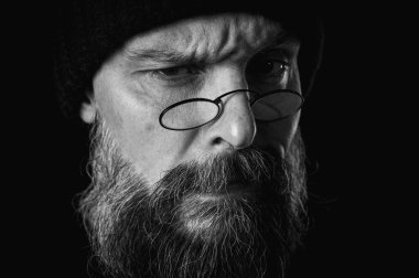 Siyah beyaz portre, gözlüklü ve örgü şapkalı kızgın sakallı adamın duygusal portresi, kameraya hırıltı, suratını buruşturma. Loş ışık, anlamlı yüz, duygusal gerilim, karanlık arka plan dramatik portre.