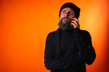 Siyah şapkalı, sakallı ve gözlüklü, parlak turuncu arka planda duran zarif olgun bir adamın portresi. Moda, kendine güven, modern görünüş.