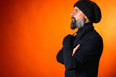 Siyah şapkalı, sakallı ve gözlüklü, parlak turuncu arka planda duran zarif olgun bir adamın portresi. Moda, kendine güven, modern görünüş.