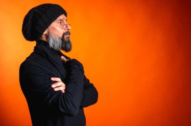 Siyah şapkalı, sakallı ve gözlüklü, parlak turuncu arka planda duran zarif olgun bir adamın portresi. Moda, kendine güven, modern görünüş.