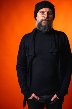 Sakallı, gözlüklü, siyah şapkalı, parlak turuncu arka planda duran şık ve olgun bir adamın portresi. Moda, kendine güven, modern görünüş.