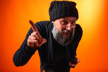 Kendinden emin, olgun bir adamın portresi elleriyle jestler yapıyor, örgü bir şapka ve turuncu bir arka planda gözlük takıyor, kameraya bakıyor. Dikkatli bak, gri sakallı. Kısıtlı portre, yazıtlar için yer.