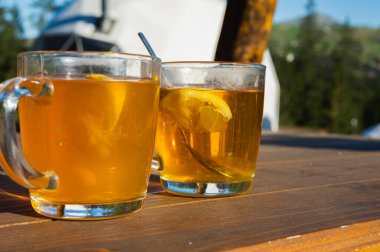 Dışarıdaki ahşap bir masada arka planda dağlar olan iki bardak sıcak limon çayı. Sıcak güneş ışığı, ferahlatıcı içecek, doğal ortam..