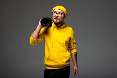 Şık, duygusal bir portre. Sarı örgü şapkalı, kazaklı, etekli ve gri arka planda kamera olan bir adam.