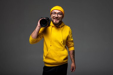 Sarı kapüşonlu ve bereli gülümseyen genç adam stüdyoda kamera tutuyor. Gri arka planda sakallı, gözlüklü ve dövmeli hippi fotoğrafçı.