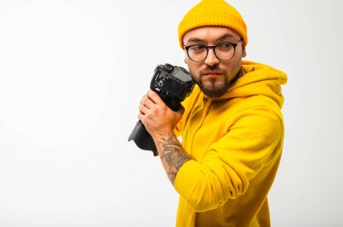 Sarı kapüşonlu ve şapkalı gülümseyen genç adam stüdyoda kamera tutuyor. Beyaz arka planda sakallı, gözlüklü ve dövmeli hippi fotoğrafçı..