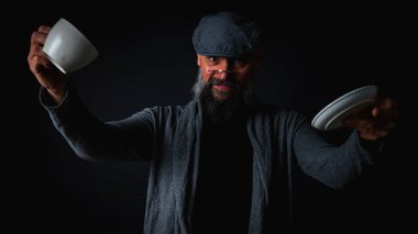 Şapkalı şık sakallı bir adam, elinde kahve fincanı ve fincan tabağı tutan koyu renk bir arka plan, duygular, el hareketleri. Dalgın yüz ifadesi, modern klasik atmosfer, sabah çay partisi..