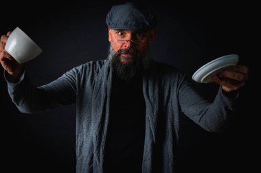 Şapkalı şık sakallı bir adam, elinde kahve fincanı ve fincan tabağı tutan koyu renk bir arka plan, duygular, el hareketleri. Dalgın yüz ifadesi, modern klasik atmosfer, sabah çay partisi..