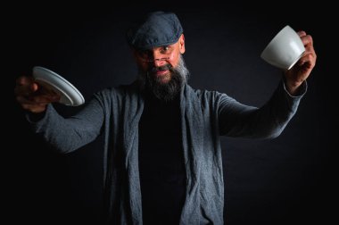 Şapkalı şık sakallı bir adam, elinde kahve fincanı ve fincan tabağı tutan koyu renk bir arka plan, duygular, el hareketleri. Dalgın yüz ifadesi, modern klasik atmosfer, sabah çay partisi..