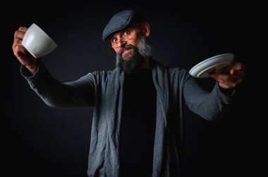 Şapkalı şık sakallı bir adam, elinde kahve fincanı ve fincan tabağı tutan koyu renk bir arka plan, duygular, el hareketleri. Dalgın yüz ifadesi, modern klasik atmosfer, sabah çay partisi..