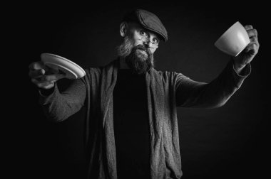 Siyah ve beyaz fotoğraf, şapkalı şık sakallı adam ve elinde kahve fincanı ve fincan tabağı tutan koyu arka planda, duygular, el hareketleri. Dalgın yüz ifadesi, modern klasik atmosfer, sabah çayı..
