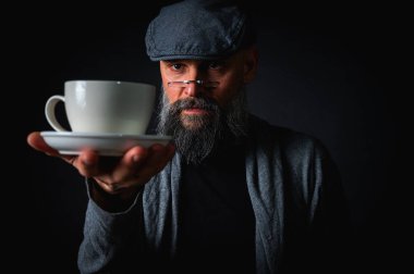 Şapkalı şık sakallı bir adam, elinde kahve fincanı ve fincan tabağı tutan koyu renk bir arka plan, duygular, el hareketleri. Dalgın yüz ifadesi, modern klasik atmosfer, sabah çay partisi..