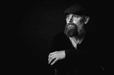 Siyah beyaz portre, siyah şapkalı ve gözlüklü duygusal sakallı adam, kameraya düşünceli bir bakış. Bastırılmış aydınlatma, anlamlı yüz, duygusal gerilim, karanlık arka plan dramatik portre.