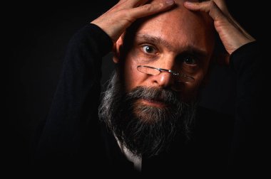 Gözlüklü ve sakallı, etkileyici ışıklandırmayla stüdyoda poz veren düşünceli olgun bir adam. Bilgeliğin kavramsal portresi, özgüven ve siyah arkaplan üzerine iç gözlem