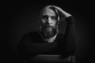 Siyah beyaz portre, siyah şapkalı ve gözlüklü duygusal sakallı adam, kameraya düşünceli bir bakış. Bastırılmış aydınlatma, anlamlı yüz, duygusal gerilim, karanlık arka plan dramatik portre.