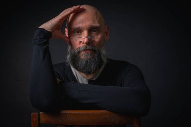 Gözlüklü ve sakallı, etkileyici ışıklandırmayla stüdyoda poz veren düşünceli olgun bir adam. Bilgeliğin kavramsal portresi, özgüven ve siyah arkaplan üzerine iç gözlem