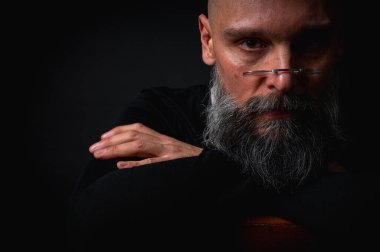 Gözlüklü ve sakallı, etkileyici ışıklandırmayla stüdyoda poz veren düşünceli olgun bir adam. Bilgeliğin kavramsal portresi, özgüven ve siyah arkaplan üzerine iç gözlem