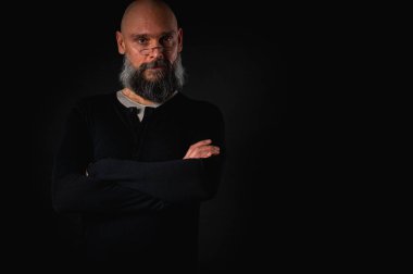 Siyah gömlekle siyah arka planda poz veren çapraz kollu kendine güvenen sakallı bir adam. Olgun hipster tarzı. Güçlü karakter ve karizmayla stüdyo portresi