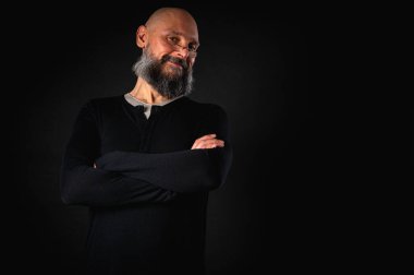 Siyah gömlekle siyah arka planda poz veren çapraz kollu kendine güvenen sakallı bir adam. Olgun hipster tarzı. Güçlü karakter ve karizmayla stüdyo portresi
