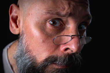 Gözlüklü kızgın sakallı adamın duygusal portresi kameraya hırıldıyor, suratını ekşitiyor. Loş ışık, anlamlı yüz, duygusal gerilim, karanlık arka plan dramatik portre.