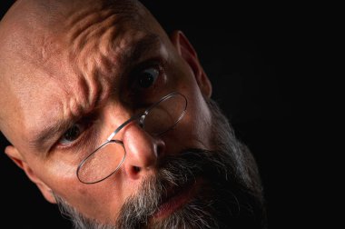 Gözlüklü kızgın sakallı adamın duygusal portresi kameraya hırıldıyor, suratını ekşitiyor. Loş ışık, anlamlı yüz, duygusal gerilim, karanlık arka plan dramatik portre.