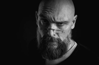 Siyah beyaz portre, gözlüklü duygusal sakallı adam, suratını buruşturup kameraya bakıyor. Loş ışık, anlamlı yüz, duygusal gerilim, karanlık arka plan dramatik portre.