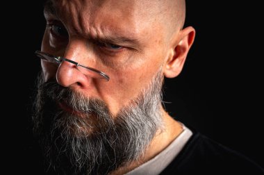 Gözlüklü kızgın sakallı adamın duygusal portresi kameraya hırıldıyor, suratını ekşitiyor. Loş ışık, anlamlı yüz, duygusal gerilim, karanlık arka plan dramatik portre.