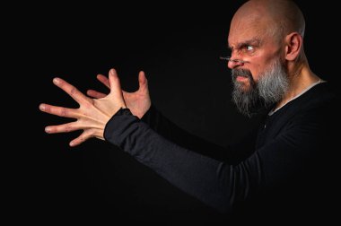 Sakallı ve gözlüklü enerjik bir adamın dramatik portresi. Duygusal olarak kenara uzanıp bir şeyler yakalamaya çalışıyor. İnce ışıklandırma, duygusal ifade, karanlık arkaplan, gizemli konsept.