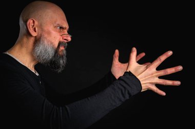 Kameraya uzanan sakallı ve gözlüklü enerjik bir adamın dramatik portresi. Elleriyle çekmeye çalışıyor. İnce ışıklandırma, duygusal ifade, karanlık arkaplan, gizemli konsept