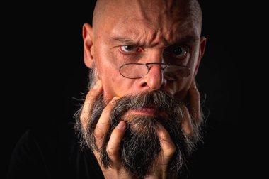 Siyah ve beyaz portre, ciddi kel, sakallı ve gözlüklü adam kameraya dikkatlice bakıyor, sakalı yüzüne yakın tutuyor. Siyah arkaplanda düşük tuşlu dramatik bir portre.