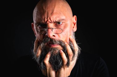 Sakallı ve gözlüklü ciddi bir kel adam kameraya dikkatlice bakıyor, elinde bir cüzdan tutuyor. Siyah arkaplanda dizginlenmiş bir şekilde dramatik bir portre.