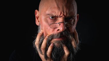 Sakallı ve gözlüklü ciddi bir kel adam kameraya dikkatlice bakıyor, elinde bir cüzdan tutuyor. Siyah arkaplanda dizginlenmiş bir şekilde dramatik bir portre.