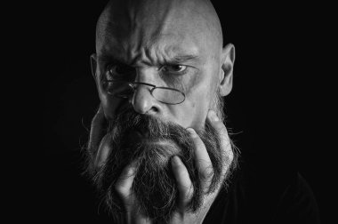 Siyah ve beyaz portre, ciddi kel, sakallı ve gözlüklü adam kameraya dikkatlice bakıyor, sakalı yüzüne yakın tutuyor. Siyah arkaplanda düşük tuşlu dramatik bir portre.