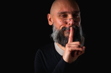 Sakallı adam sessiz hareketler yapıyor. Parmakları dudaklarında. Gizemli gizli konsept. Dramatik ışıklandırma ve ciddi ifade ile siyah arkaplanda portre