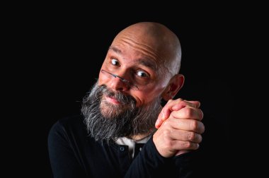 Gri sakallı, gözlüklü, kurnaz görünümlü kel bir adamın portresi. Karanlık arka plan. Yaramaz ya da entrikacı ifade. Komplo, iğneleme ya da ironik mizah anlayışı