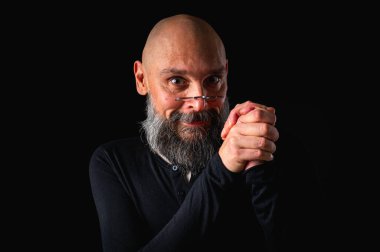 Gri sakallı, gözlüklü, kurnaz görünümlü kel bir adamın portresi. Karanlık arka plan. Yaramaz ya da entrikacı ifade. Komplo, iğneleme ya da ironik mizah anlayışı